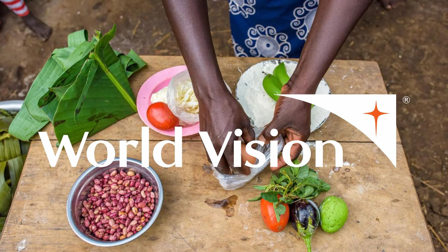 A person prepares food on a wooden table with vegetables, beans, and leaves, viewed from above. The World Vision logo is overlaid on the image.
